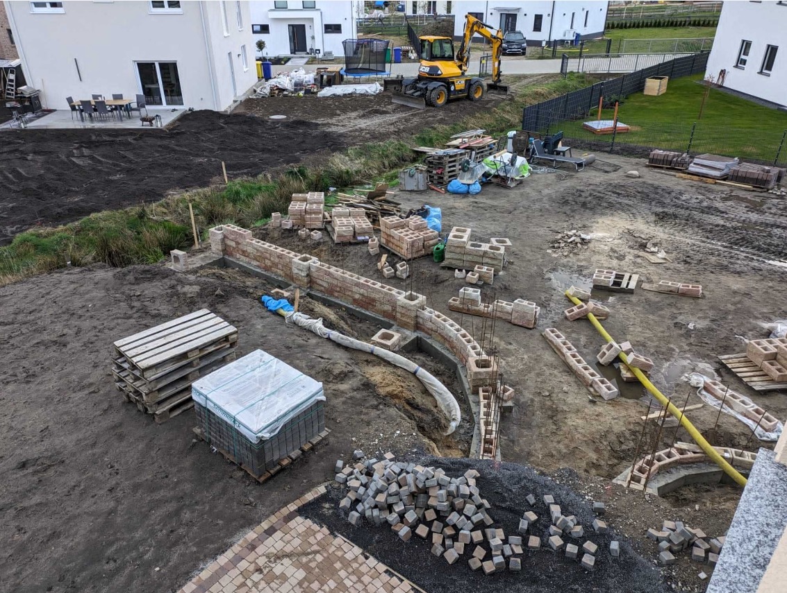 Plac budowy z wykopem pod fundamenty i częściowo wzniesionym murem z bloczków, palety z materiałami budowlanymi, koparka w tle.