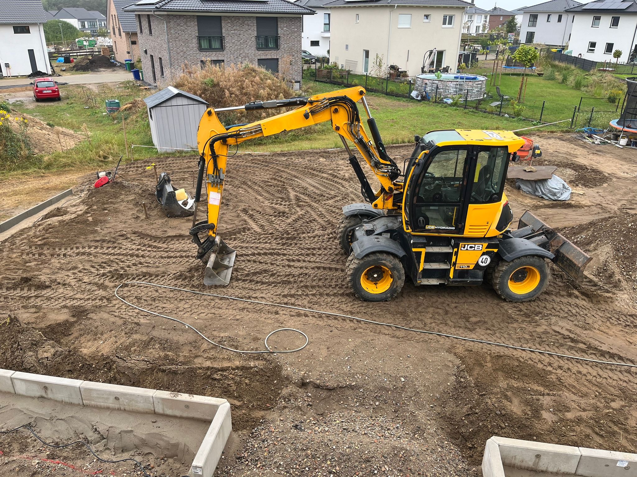 Żółta koparka JCB na placu budowy, przygotowany teren pod kostkę brukową, widoczne obrzeża. W tle domy jednorodzinne i trawnik.