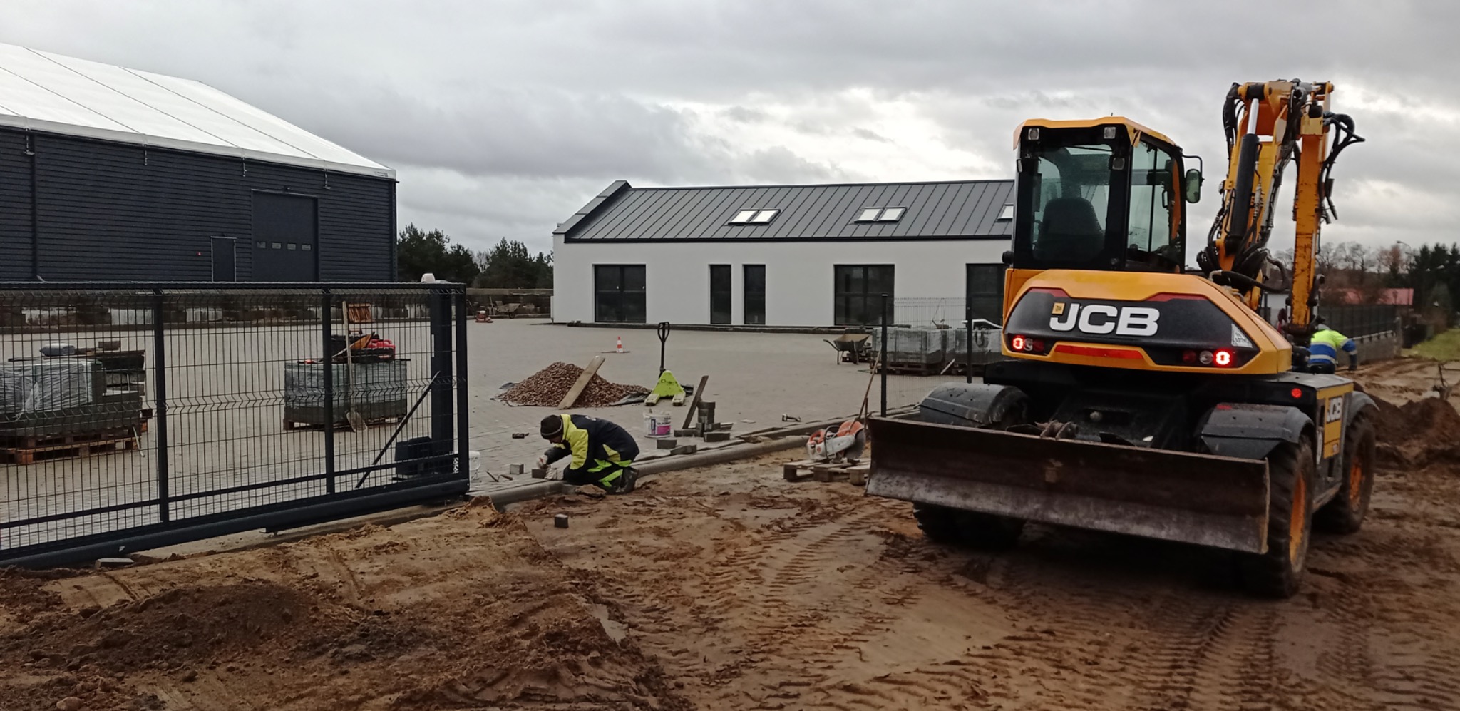 Pracownik w trakcie układania obrzeży z kostki przy wjeździe na posesję. W tle budynki i maszyna JCB. Etap prac brukarskich.