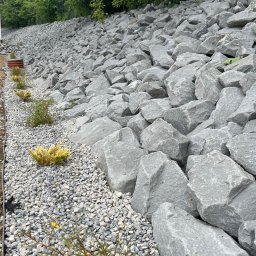 Centrum ogrodnicze pod brzoza - Skarpa umocniona dużymi, szarymi kamieniami. U podnóża wysypany drobny żwir, z posadzonymi niskimi krzewami iglastymi. W tle widoczne drzewa.