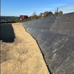 Centrum ogrodnicze pod brzoza - Przygotowanie terenu pod trawnik z rolki: wyrównany teren z widocznymi śladami stóp i rozłożona czarna geowłóknina z czerwoną siatką na zboczu.