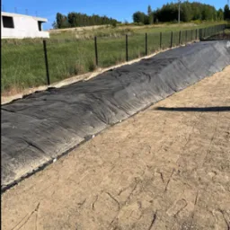 Przygotowanie podłoża pod trawnik z rolki: czarna geowłóknina rozłożona na ziemi, widoczne odciski stóp. W tle zielona trawa i ogrodzenie.