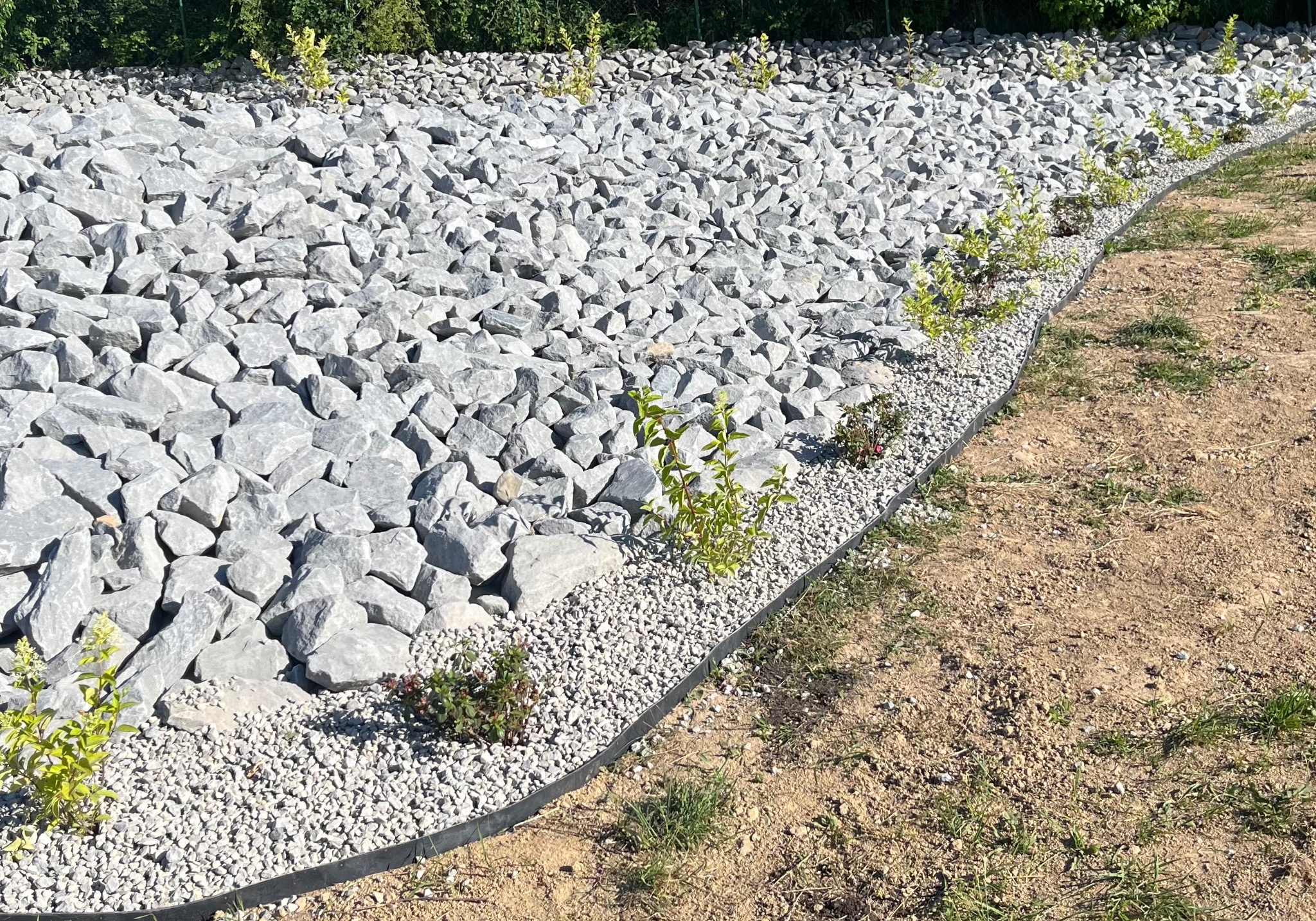 Aranżacja terenu z szarymi kamieniami i drobnym żwirem, obrzeża z tworzywa, świeżo posadzone krzewy. Widok z góry.