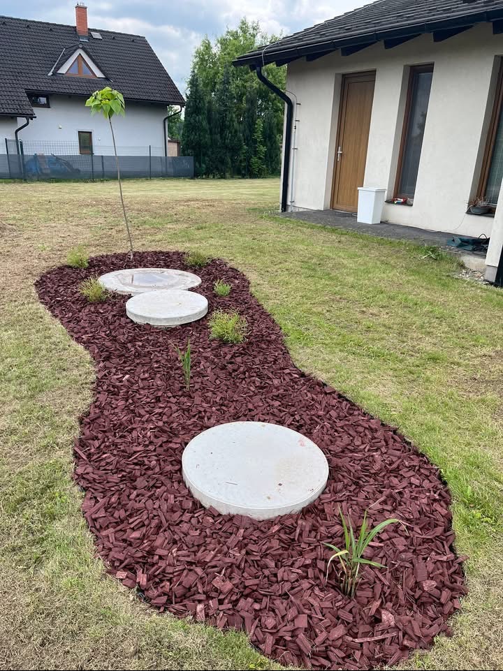 Aranżacja ogrodu z bordową korą wokół betonowych kręgów i młodej katalpy. Zielony trawnik i budynek w tle. Kora tworzy ścieżkę.