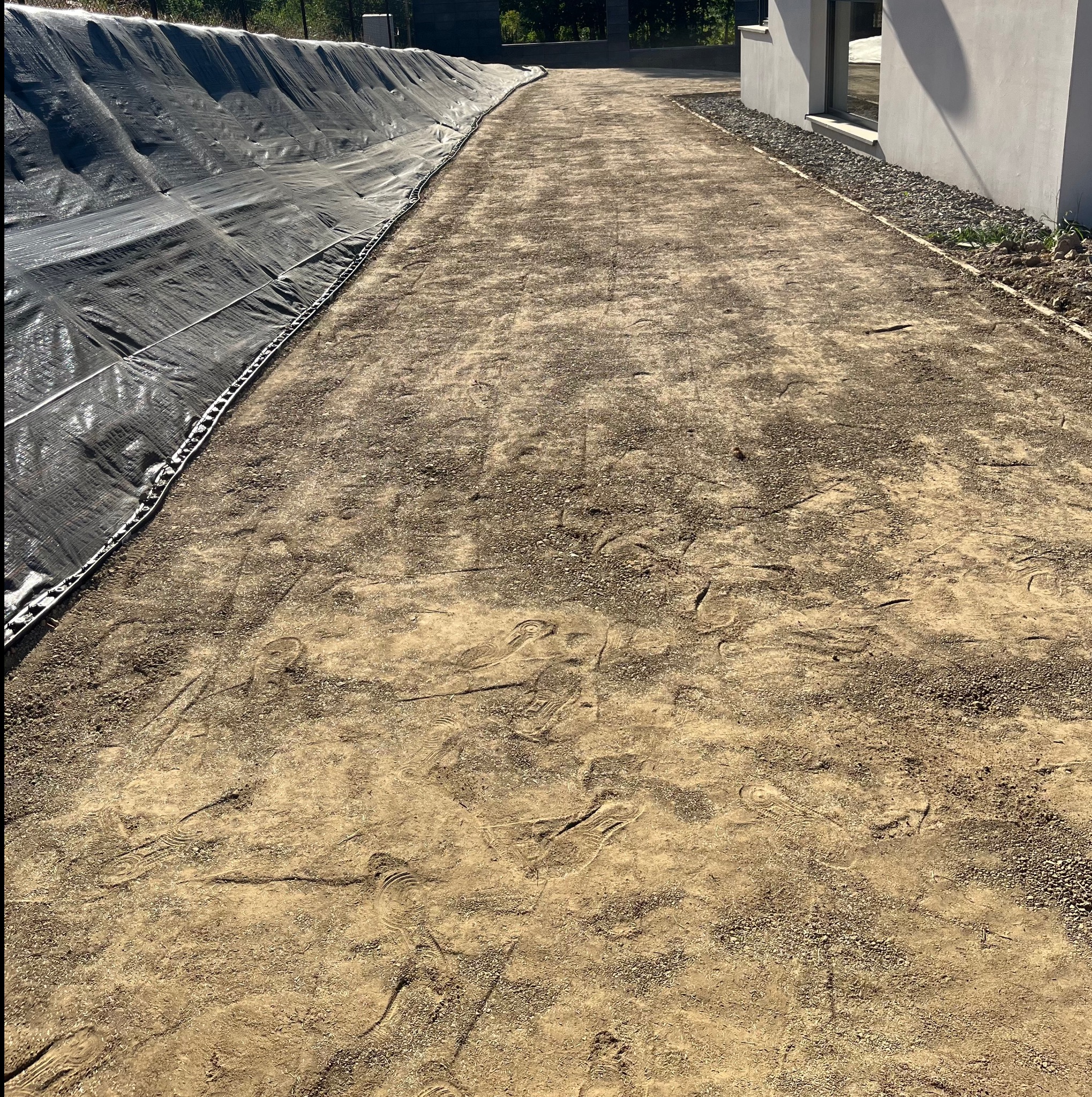 Przygotowany teren pod trawnik z widocznymi śladami butów i czarną folią ochronną przy krawędzi. Widoczny fragment budynku z szarą elewacją i okno.