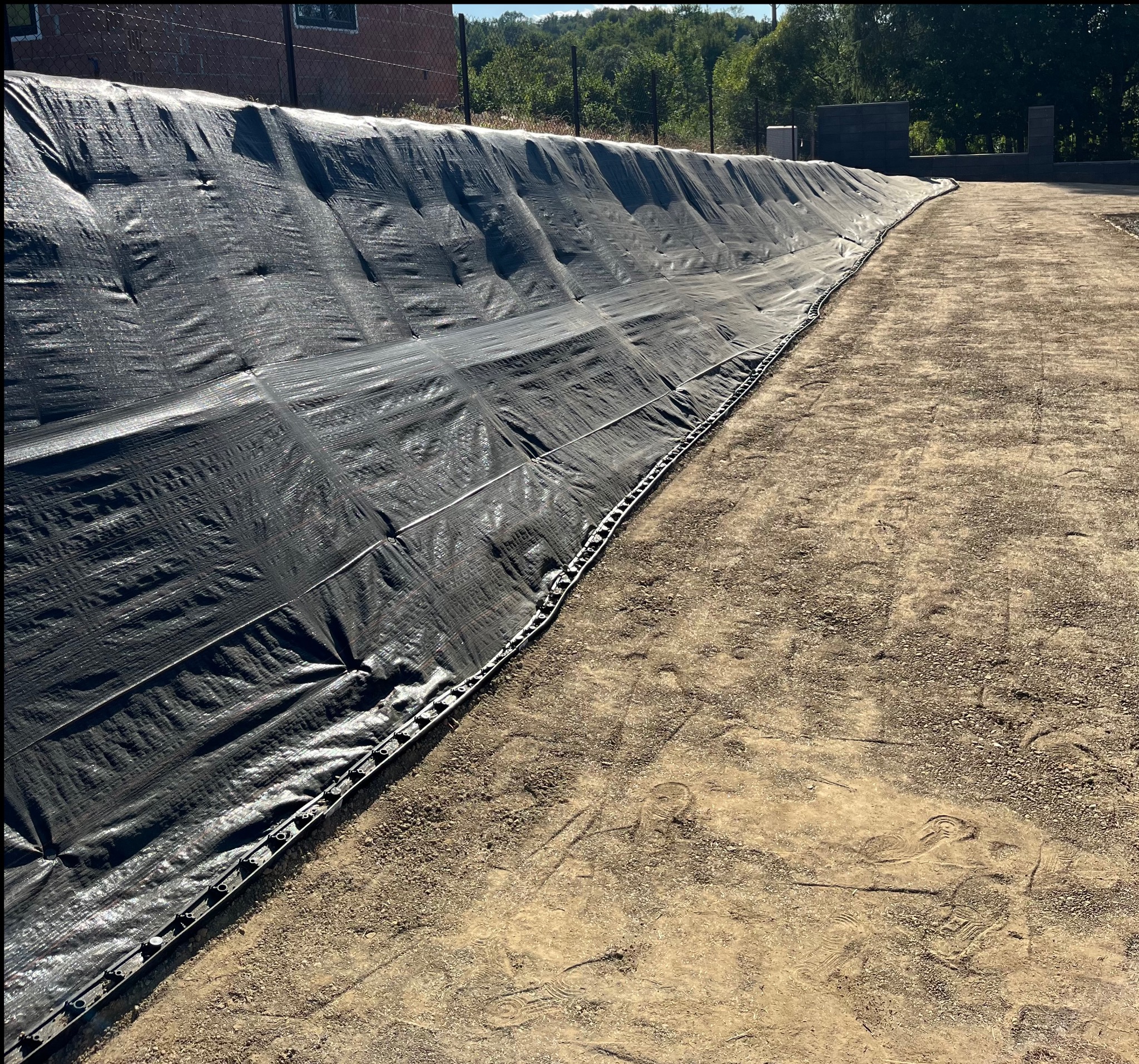 Czarna folia zabezpieczająca skarpę przed erozją, umocniona systemem kotwiącym. Tło: drzewa i fragment ogrodzenia. Widoczny przygotowany grunt.