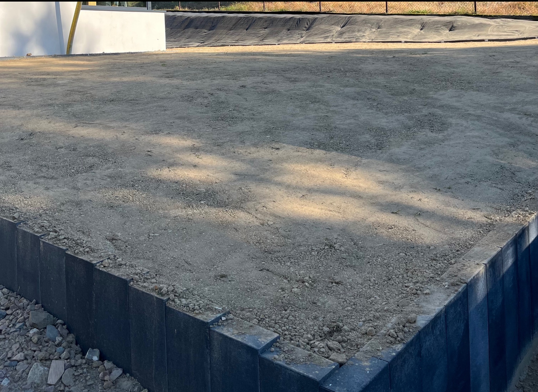 Przygotowany teren pod trawnik z widocznymi obrzeżami i membraną w tle. Ziemia wyrównana, gotowa do dalszych prac.