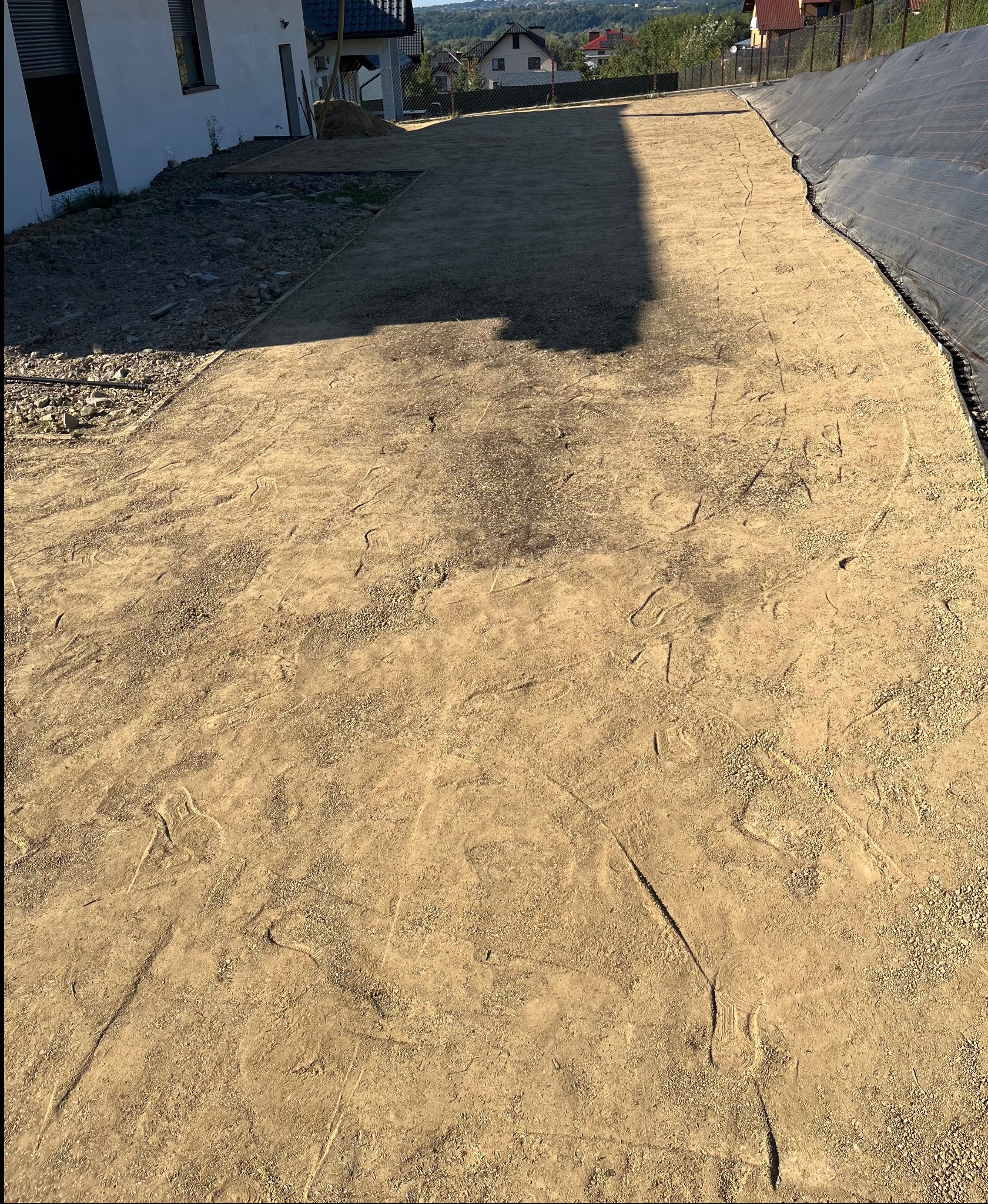 Przygotowany teren pod trawnik z rolki, widoczne ślady stóp na wyrównanej powierzchni z piasku i ziemi. W tle dom i fragment czarnej agrowłókniny.