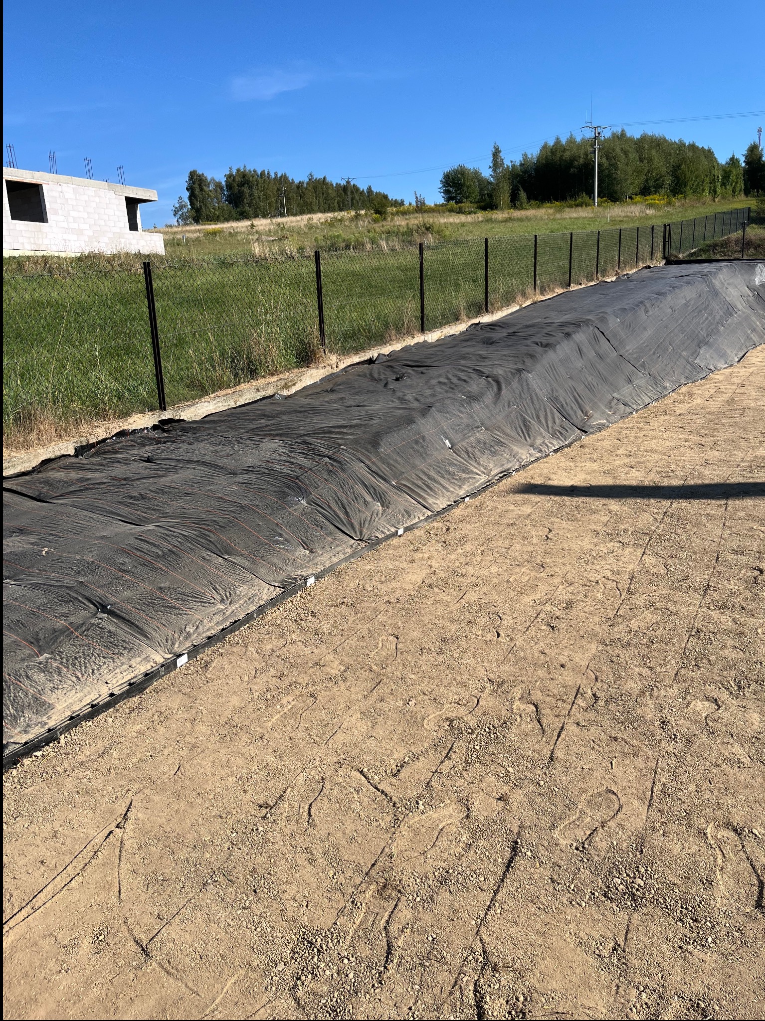 Przygotowanie podłoża pod trawnik z rolki: czarna geowłóknina rozłożona na ziemi, widoczne odciski stóp. W tle zielona trawa i ogrodzenie.