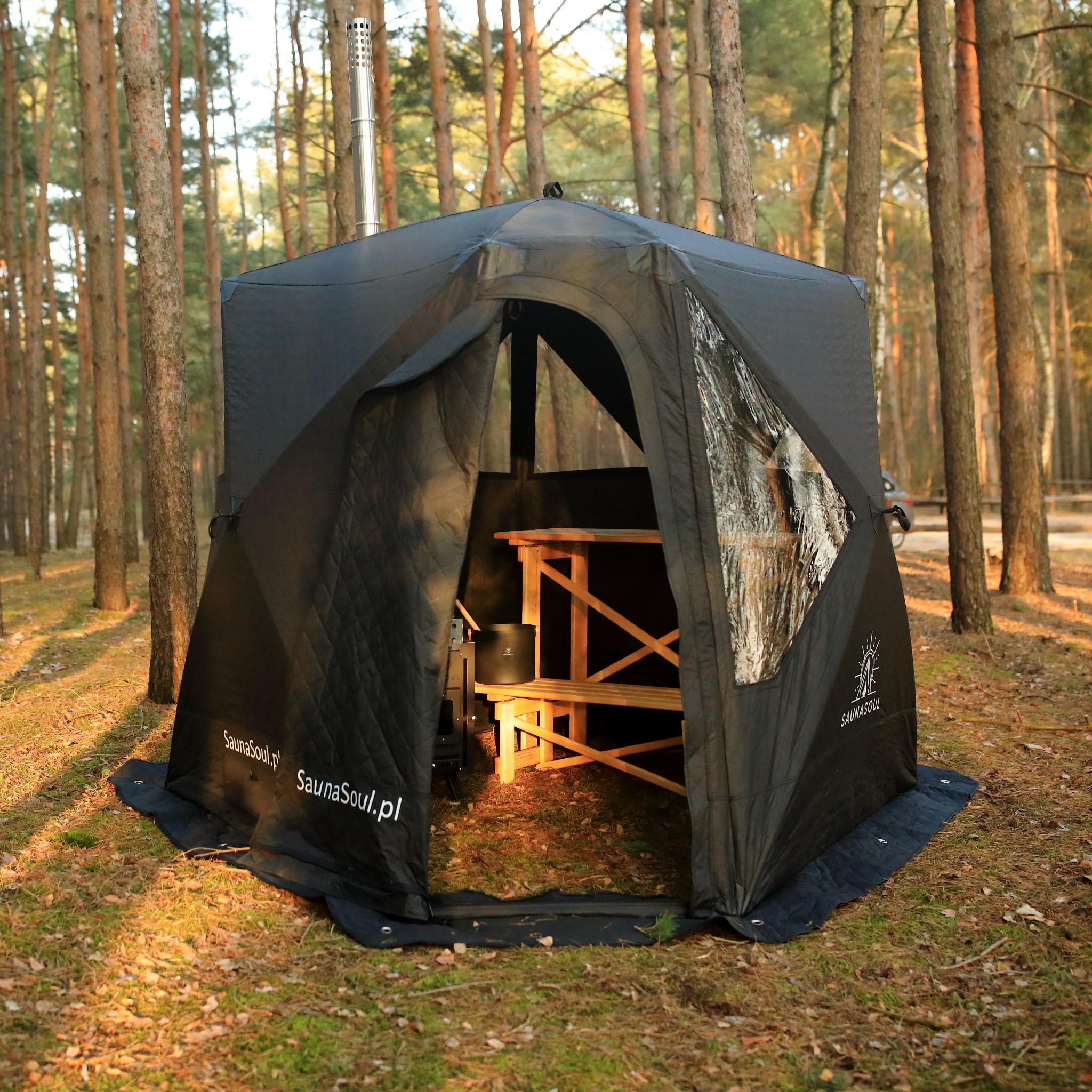 Czarna, rozkładana sauna mobilna w lesie, z widocznym piecem i drewnianą ławką wewnątrz. Na ścianie logo Saunasoul.pl. Metalowy komin wystaje z dachu.
