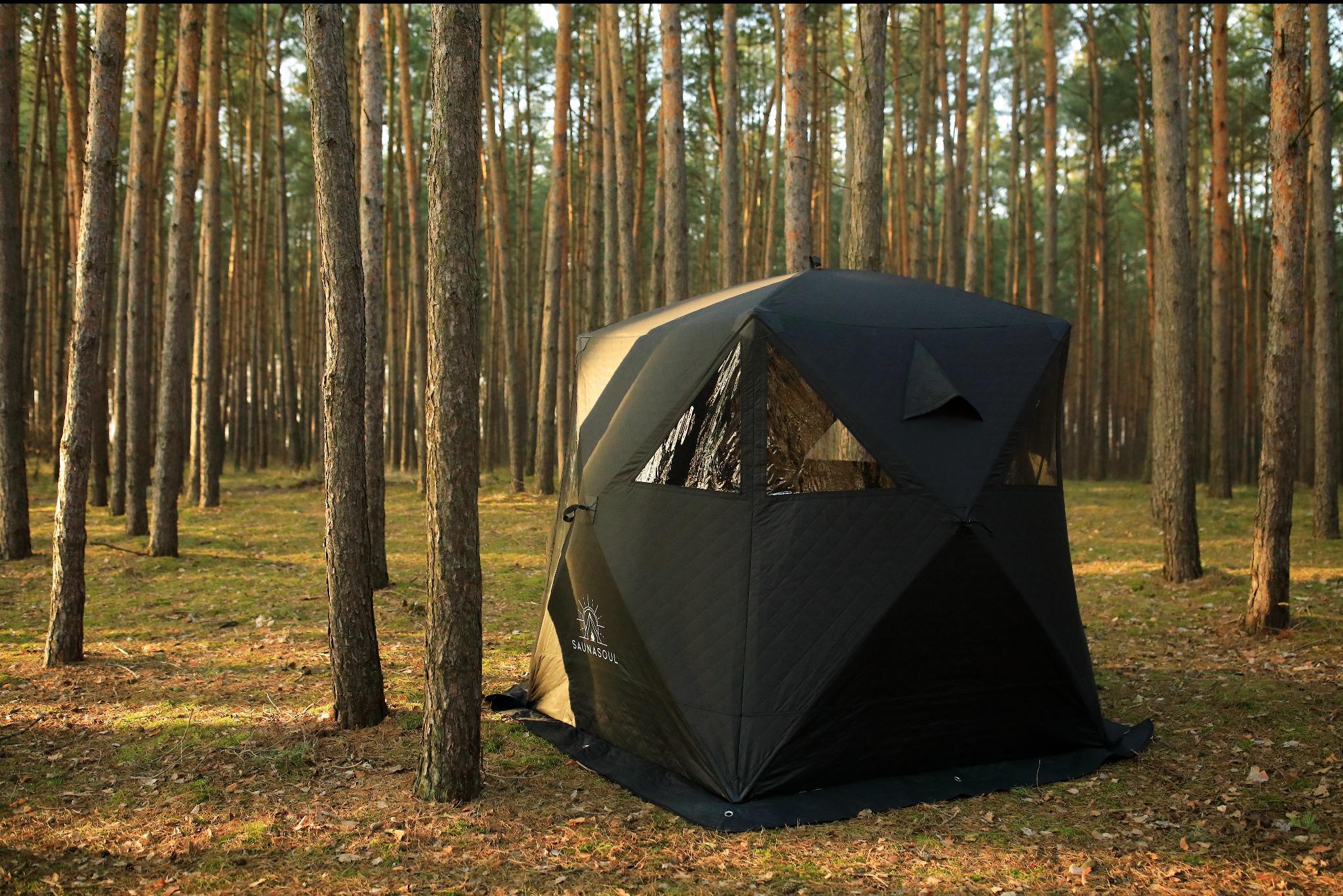 Czarna, mobilna sauna w kształcie namiotu z oknami, ustawiona w lesie sosnowym. Widoczne logo 'SAUNASOUL'.