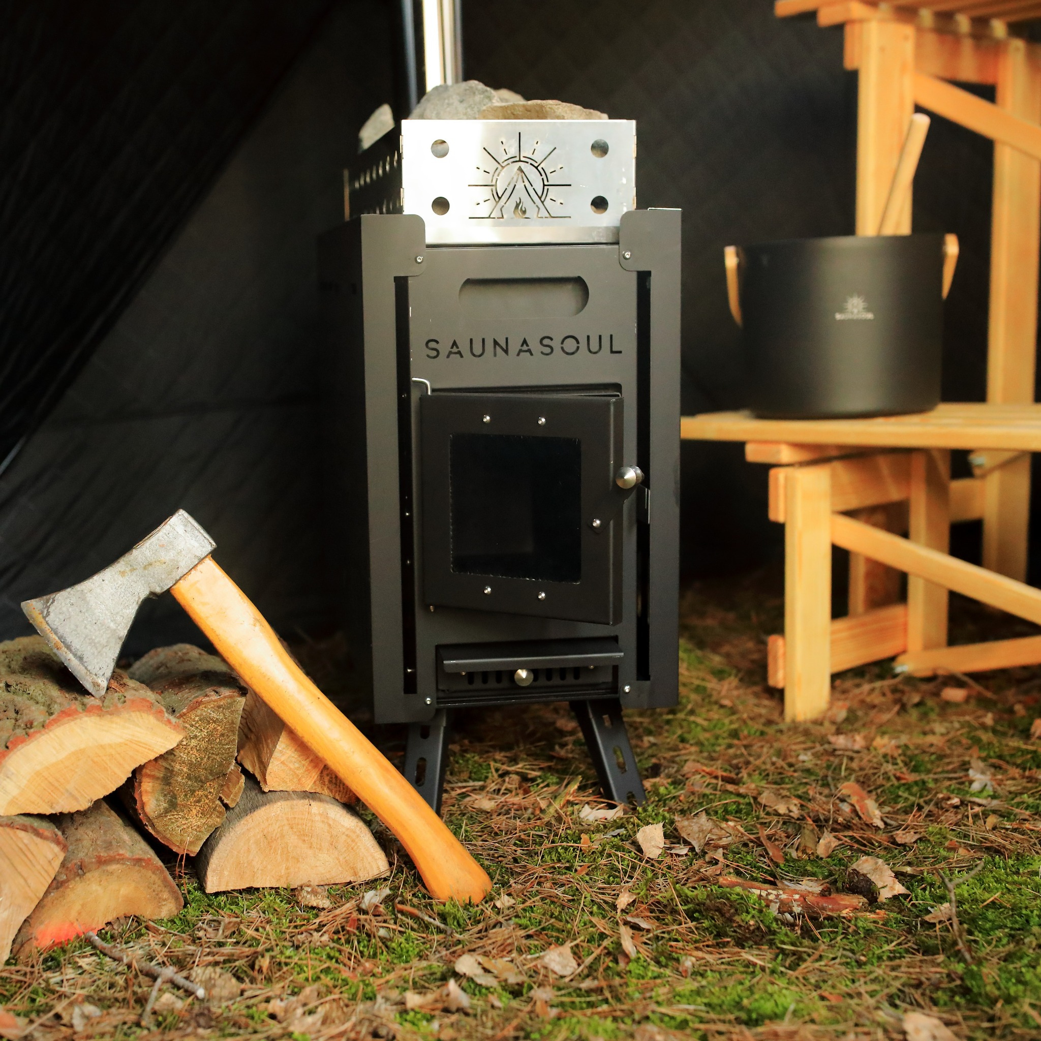 Czarny piec do sauny Saunasoul z drewnem i siekierą na zewnątrz, obok drewniany stołek z wiaderkiem. Widok na wnętrze sauny.
