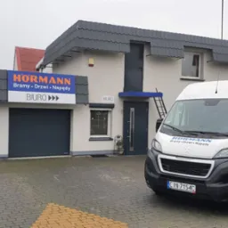 Siedziba firmy Hörmann z bramą garażową, drzwiami i napędami, z zaparkowanym samochodem dostawczym z logo firmy na tle biura. Widoczny szyld z nazwą firmy.