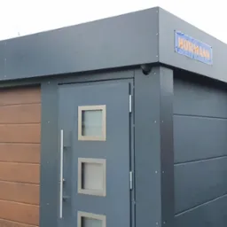 Garaż z brązową bramą segmentową i szarymi drzwiami bocznymi z trzema kwadratowymi okienkami, całość pod szyldem Hormann, na tle zabudowań.