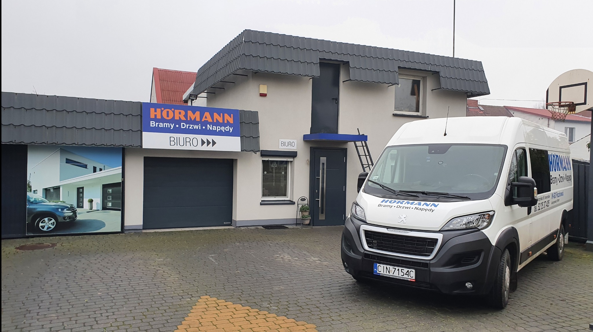 Siedziba firmy Hörmann z bramą garażową, drzwiami i napędami, z zaparkowanym samochodem dostawczym z logo firmy na tle biura. Widoczny szyld z nazwą firmy.