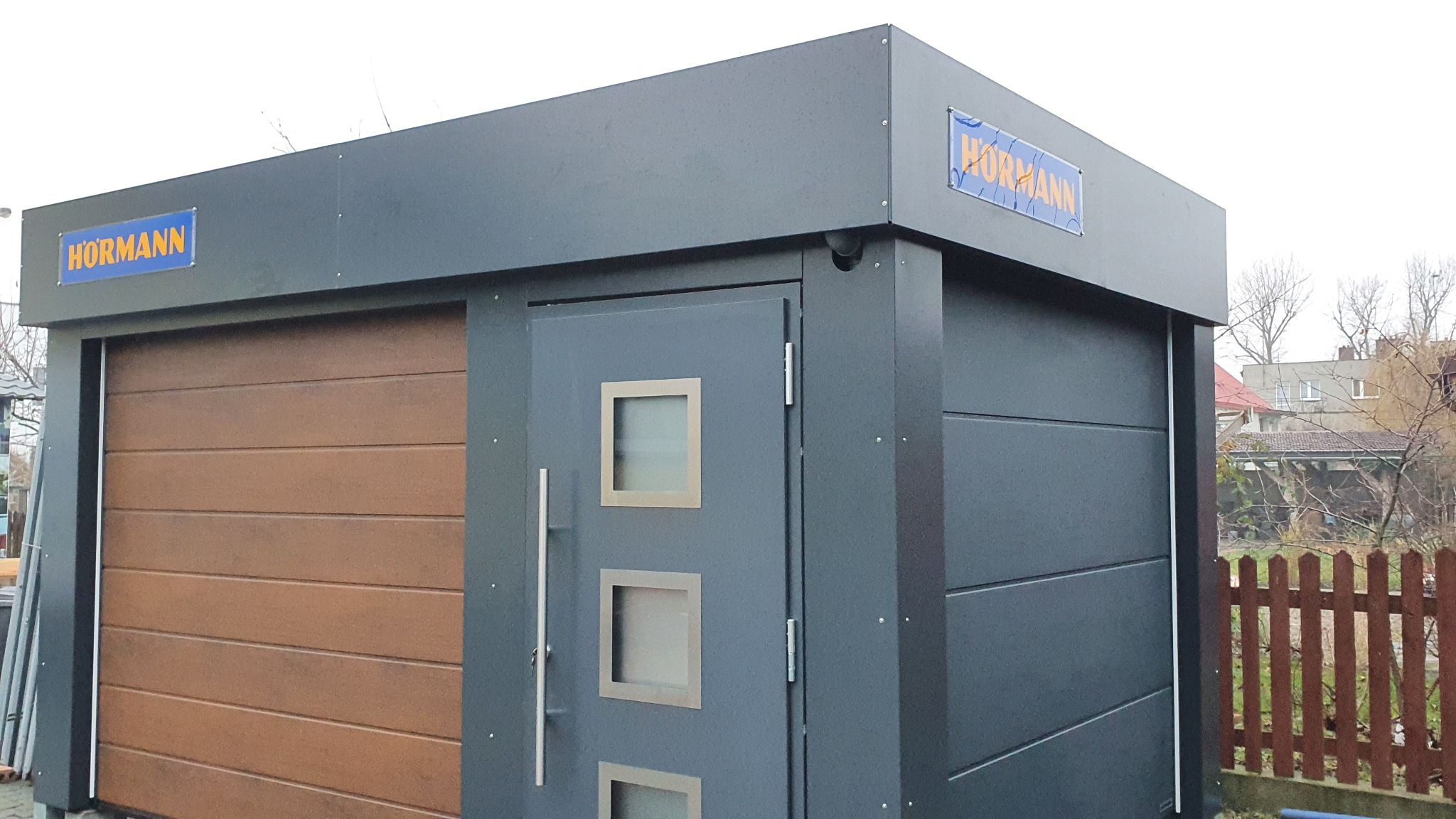 Garaż z brązową bramą segmentową i szarymi drzwiami bocznymi z trzema kwadratowymi okienkami, całość pod szyldem Hormann, na tle zabudowań.