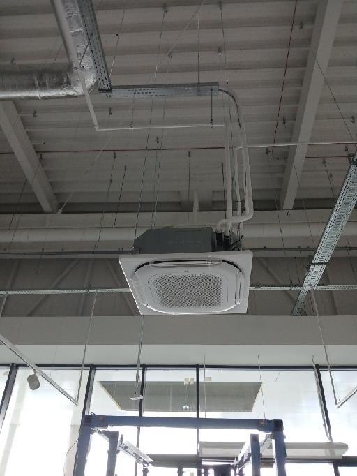 Kasetonowa jednostka klimatyzacji zamontowana pod sufitem w pomieszczeniu biurowym. Widoczne rury i kable podłączeniowe oraz metalowe koryta kablowe.