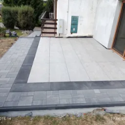 Nowoczesny taras z jasnych płyt i obramowania z szarej kostki, częściowo ukończony, z widocznymi elementami montażowymi i fragmentem domu w tle.