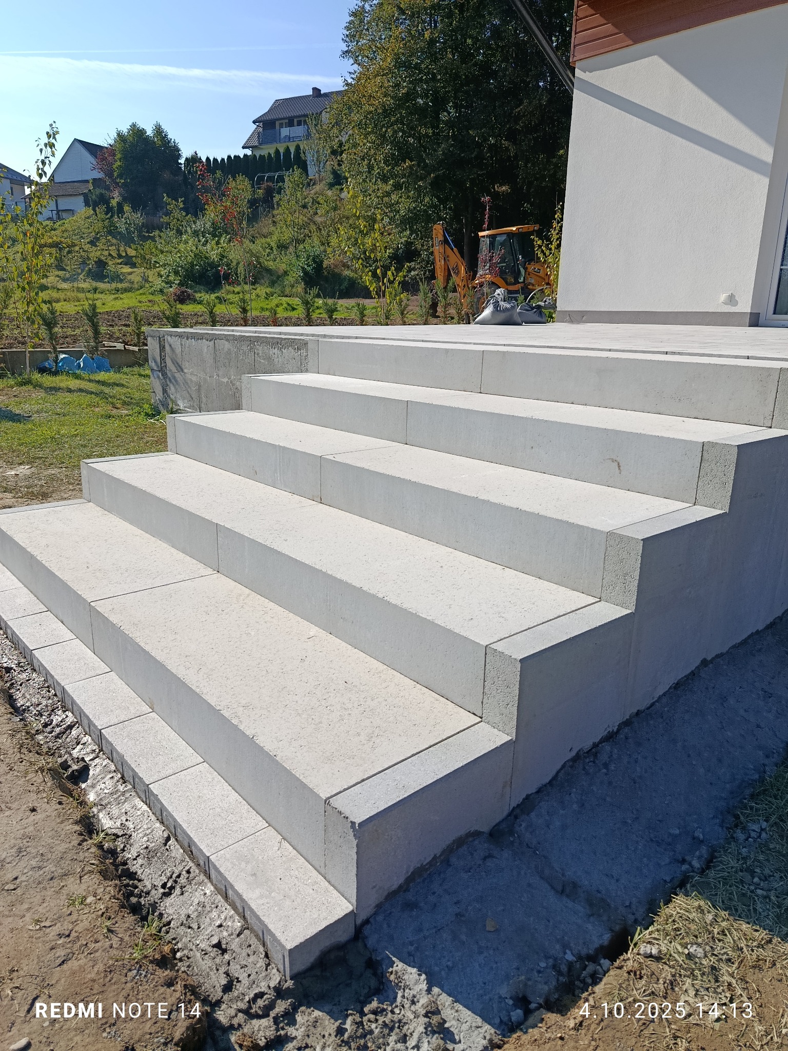 Nowe, jasnoszare betonowe schody zewnętrzne prowadzące do budynku, z widocznym wykopem i sprzętem budowlanym w tle, w słoneczny dzień.