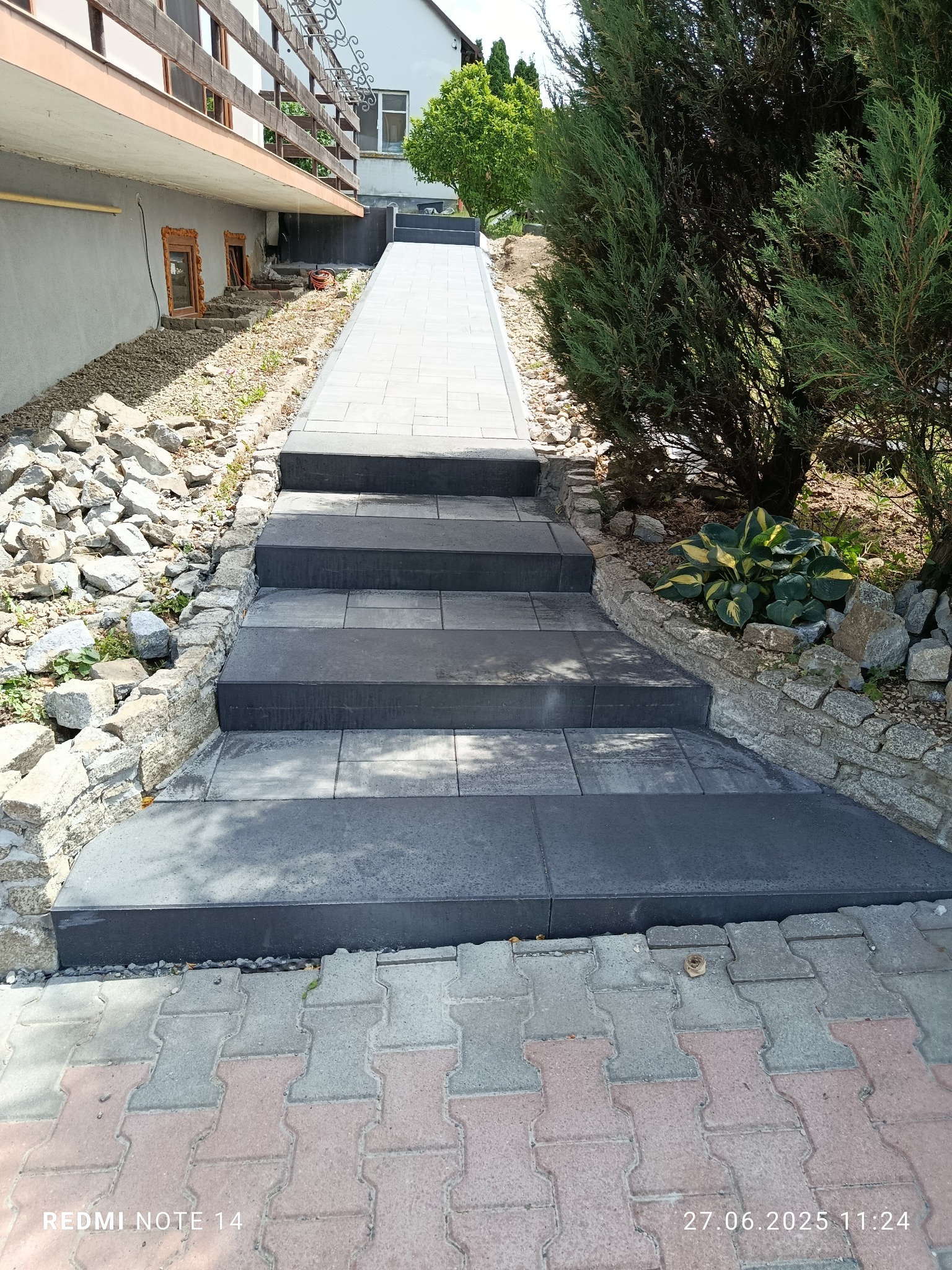 Schody z ciemnej kostki brukowej prowadzące w górę, obłożone kamieniami, z roślinnością po bokach. Widok z dołu, w tle budynek.