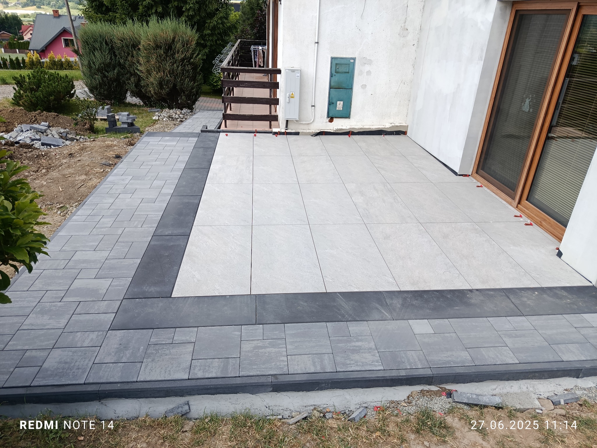 Nowoczesny taras z jasnych płyt i obramowania z szarej kostki, częściowo ukończony, z widocznymi elementami montażowymi i fragmentem domu w tle.