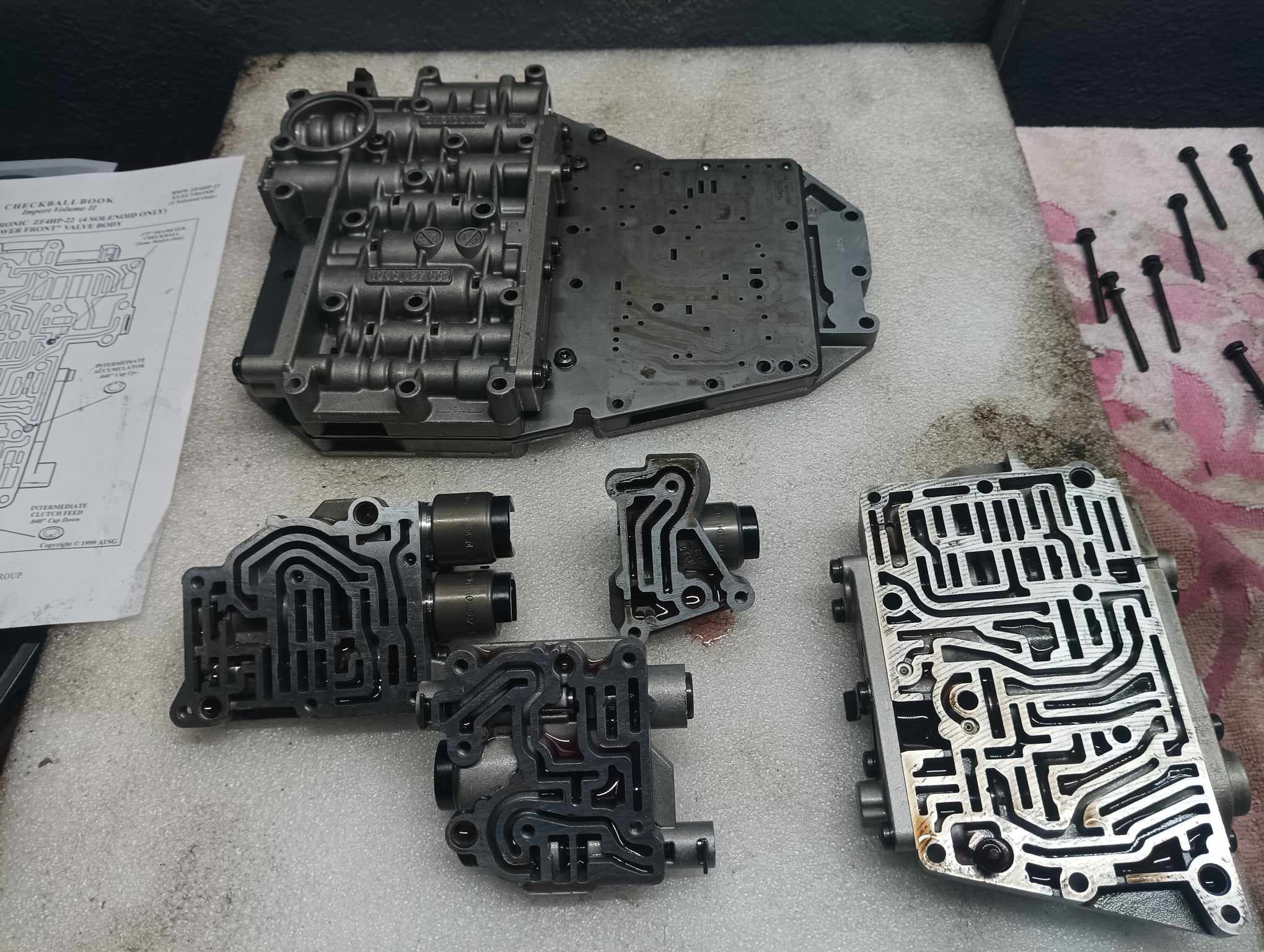 Rozłożony korpus zaworów skrzyni biegów na warsztatowym stole z instrukcją. Widoczne elementy składowe i śruby. Precyzyjna naprawa automatycznej skrzyni biegów.