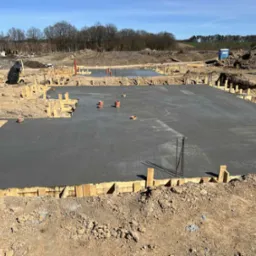 Zalany betonowy fundament pod budowę domu, z drewnianym szalunkiem i wystającymi rurami wentylacyjnymi. Widok na plac budowy w słoneczny dzień.