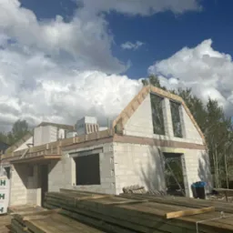 Budowa domu z białych bloczków H+H w trakcie realizacji, widoczne drewniane elementy konstrukcyjne, niebo z chmurami w tle. Częściowo zrealizowany budynek w stanie surowym.