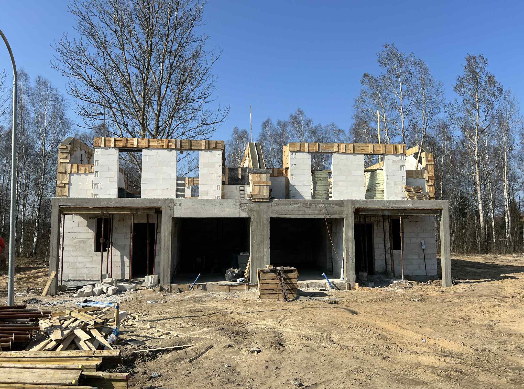 Budowa bliźniaka z betonu komórkowego: surowe ściany, drewniane szalunki okienne, betonowy strop nad garażami. Widoczne materiały budowlane i drzewa w tle.