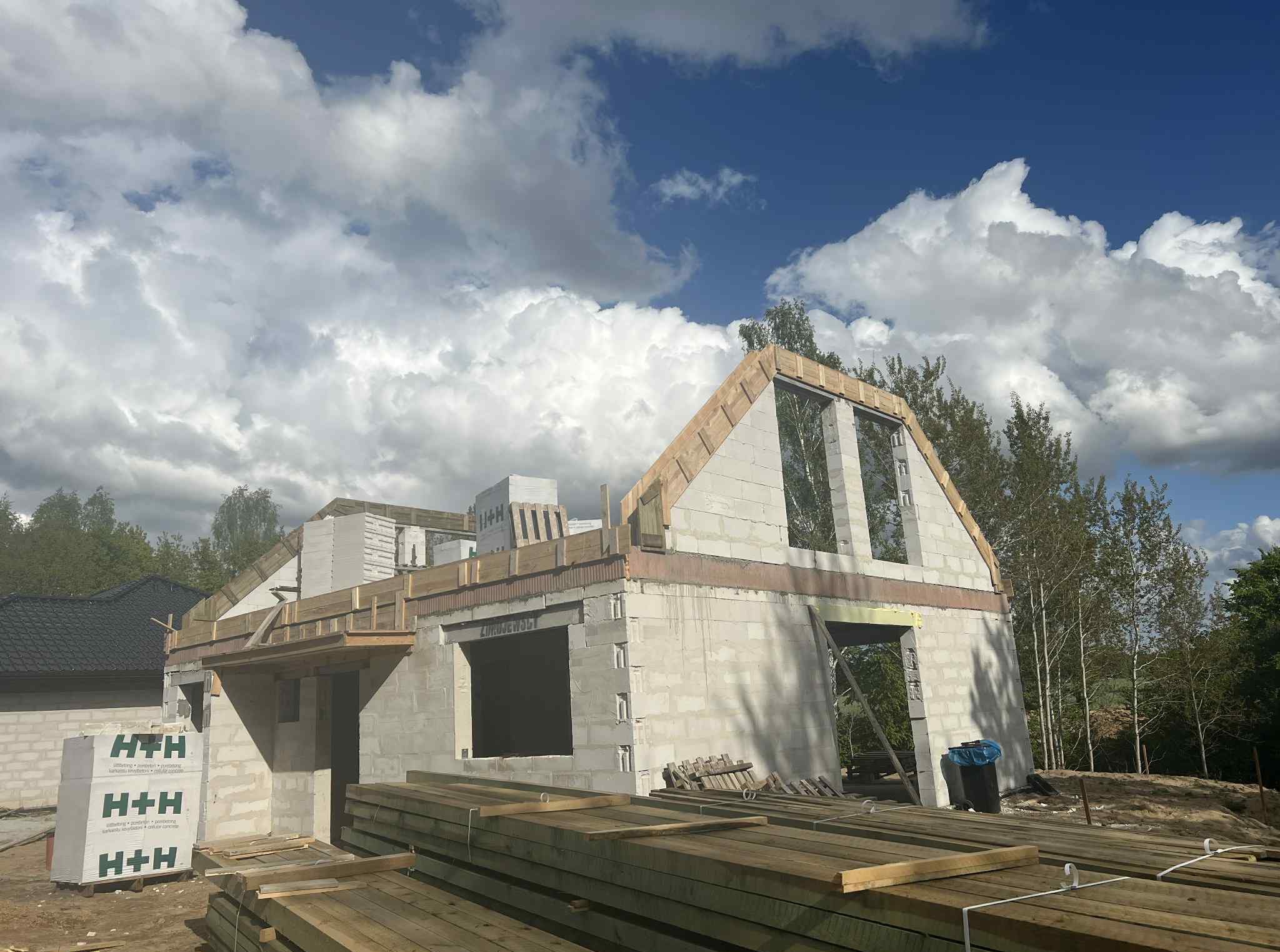 Budowa domu z białych bloczków H+H w trakcie realizacji, widoczne drewniane elementy konstrukcyjne, niebo z chmurami w tle. Częściowo zrealizowany budynek w stanie surowym.
