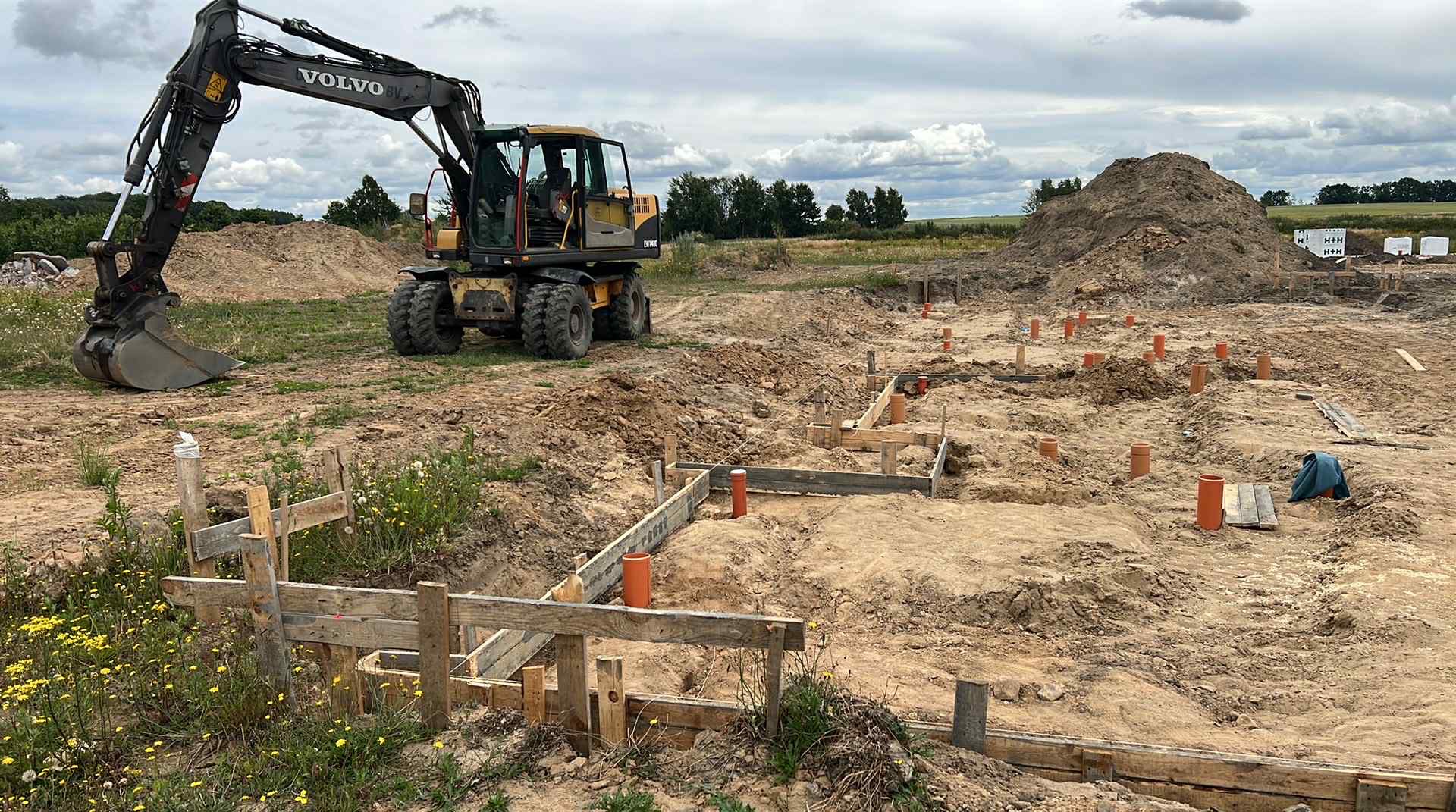 Fundamenty domu w trakcie budowy z widocznym szalunkiem, rurami i koparką Volvo w tle. Prace ziemne w toku, przygotowanie pod wylewkę.