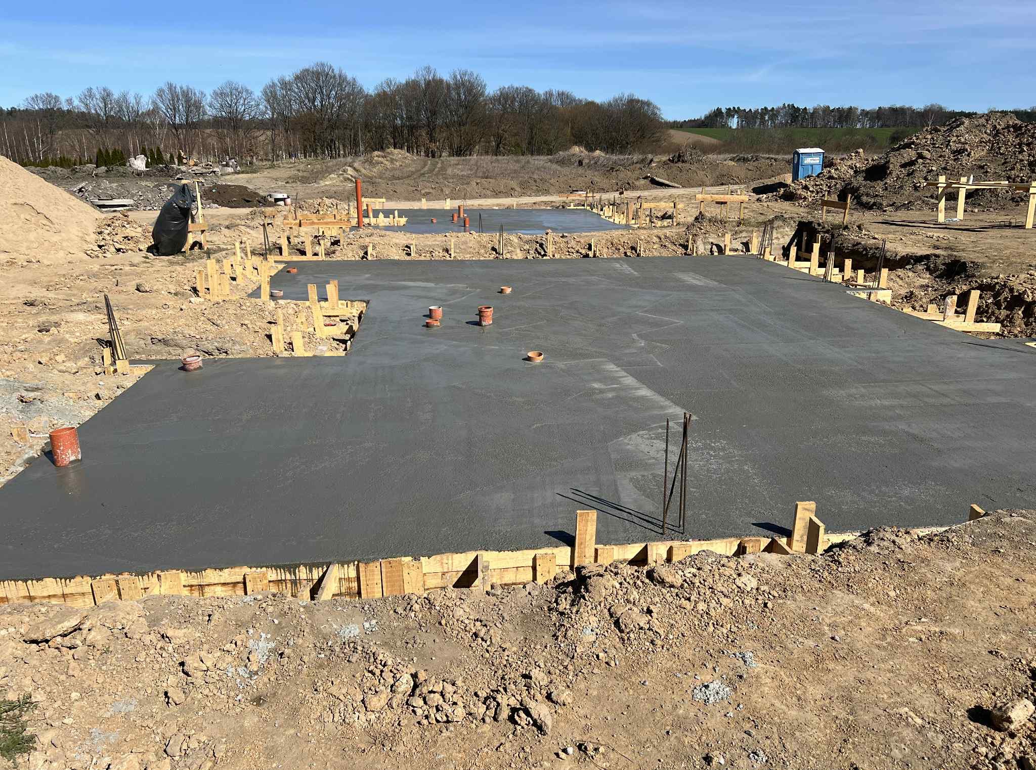 Zalane betonem fundamenty domu jednorodzinnego, szalunek z desek, wystające rury kanalizacyjne. Widok z poziomu gruntu, w tle drzewa.