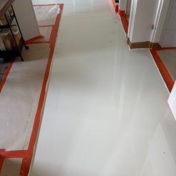 EPOXYGARAGE SPÓŁKA Z OGRANICZONĄ ODPOWIEDZIALNOŚCIĄ - Jasna, lśniąca posadzka z widocznymi odbiciami światła, świeżo położona w nowoczesnym wnętrzu. Pomarańczowa taśma maskująca zabezpiecza ściany i futryny drzwiowe.