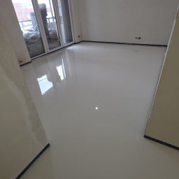 EPOXYGARAGE SPÓŁKA Z OGRANICZONĄ ODPOWIEDZIALNOŚCIĄ - Lśniąca, jasnoszara posadzka żywiczna w nowo wykończonym pokoju z dużymi oknami balkonowymi, odbijająca światło i tymczasową żarówkę. Widoczne taśmy ochronne.