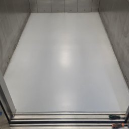 EPOXYGARAGE SPÓŁKA Z OGRANICZONĄ ODPOWIEDZIALNOŚCIĄ - Wnętrze kabiny windy z lśniącą, jasnoszarą, gładką powierzchnią podłogi. Ściany wykończone szczotkowanymi panelami metalowymi. Przy wejściu widoczne narzędzia i taśma malarska.