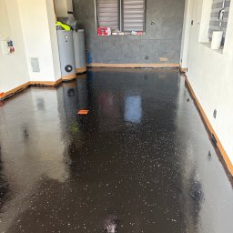 EPOXYGARAGE SPÓŁKA Z OGRANICZONĄ ODPOWIEDZIALNOŚCIĄ - Błyszcząca, czarna posadzka z białymi drobinkami, odbijająca światło, w pomieszczeniu gospodarczym z szarymi płytkami na ścianie, dwoma bojlerami i białymi ścianami. Widoczne okno z żaluzjami.