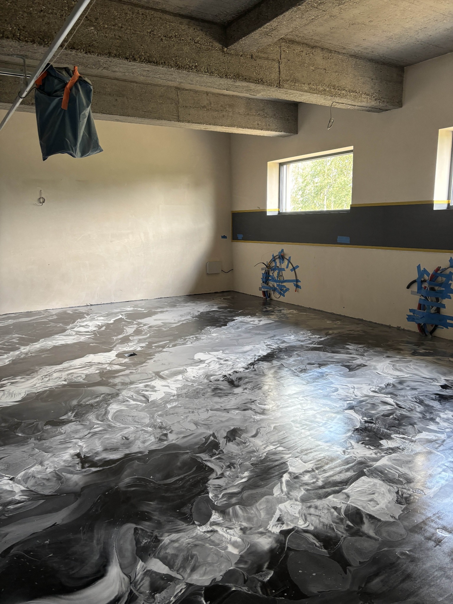 Błyszcząca, szara posadzka żywiczna z efektem marmuru w surowym wnętrzu. Widoczne instalacje zabezpieczone taśmą, okna i betonowy strop. Worki wiszą pod sufitem.