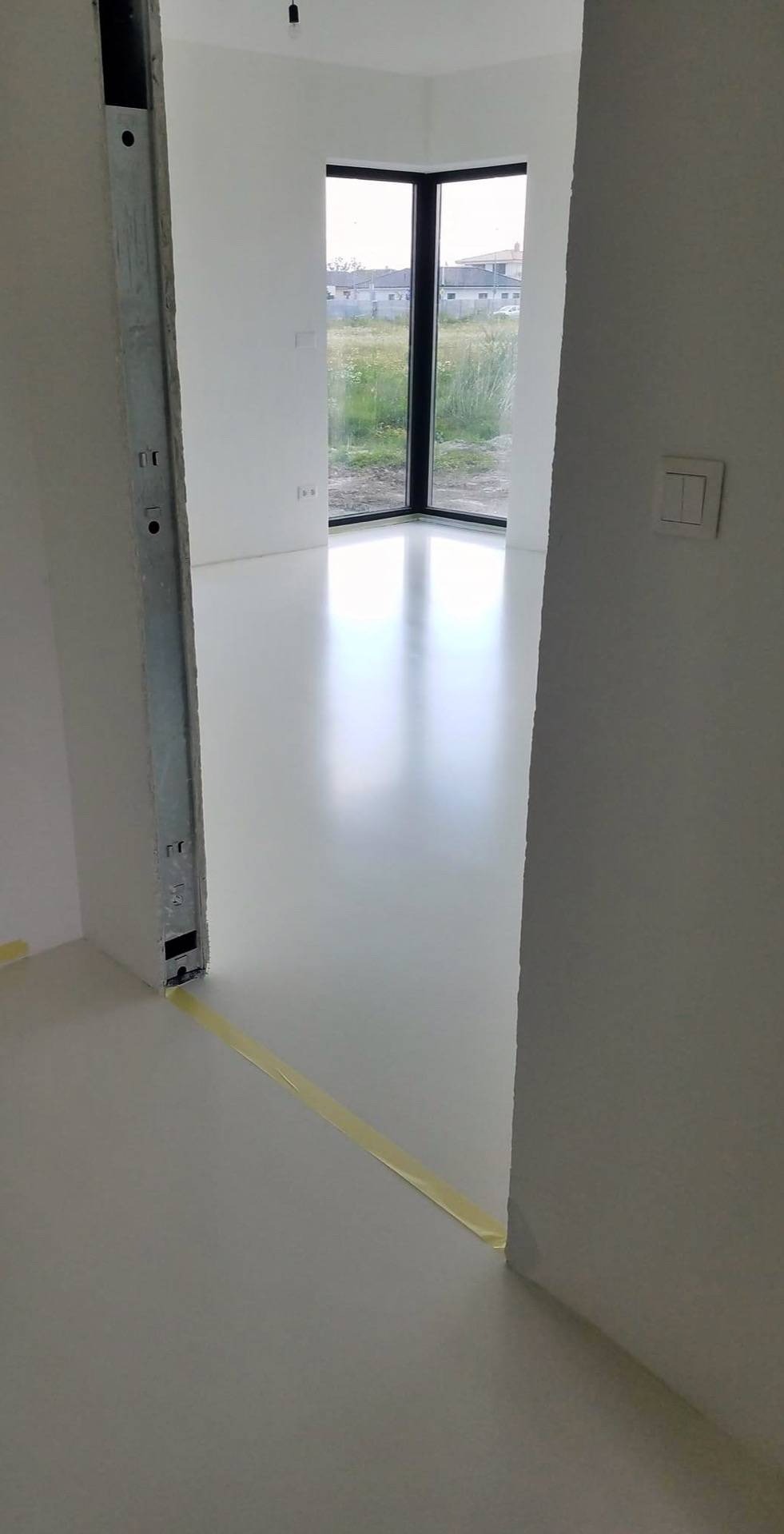 Biała, błyszcząca posadzka w jasnym pokoju z dużym oknem narożnym. Widoczny fragment metalowej futryny i żółta taśma zabezpieczająca przy podłodze. Minimalistyczny design.
