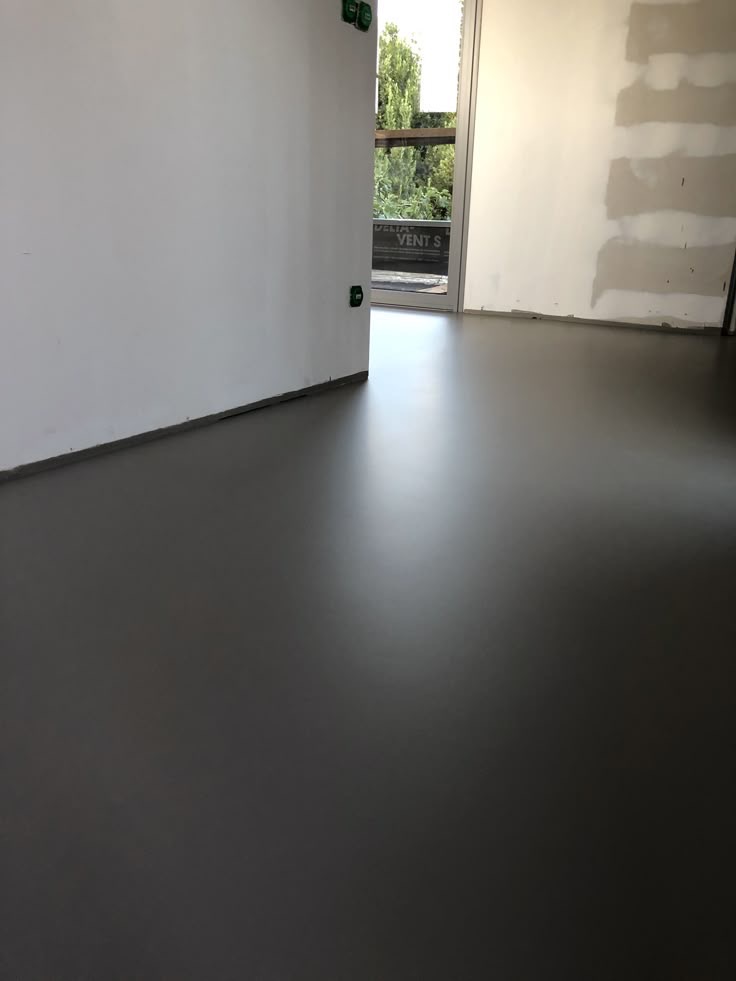 Szara, gładka posadzka w nowym pomieszczeniu z białymi ścianami i oknem. Widoczne zielone gniazdka elektryczne i fragment ściany przygotowanej do malowania.