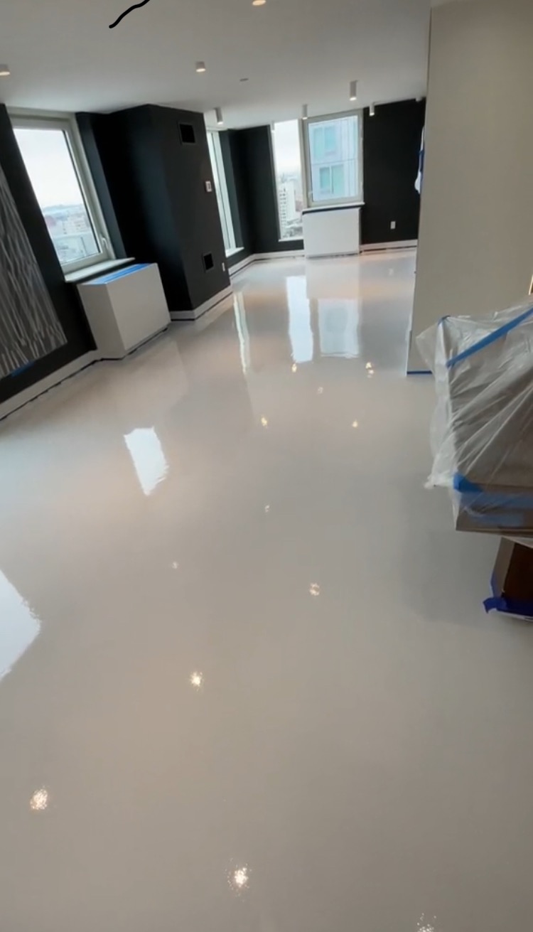 Błyszcząca, biała posadzka w nowoczesnym wnętrzu apartamentu z widokiem na miasto. Meble zabezpieczone folią malarską. Minimalistyczny design, czarne ściany.