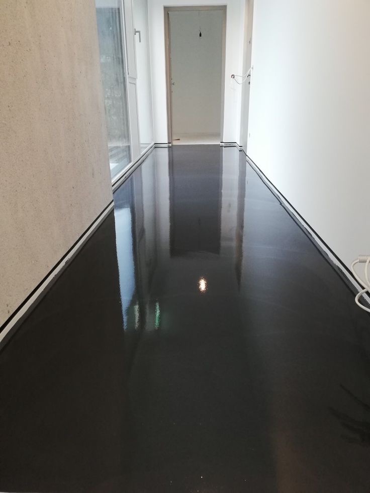 Błyszcząca, ciemna posadzka w korytarzu, z widocznym odbiciem światła. Ściany białe, z listwami przypodłogowymi. Widoczne drzwi w głębi i fragment grzejnika.