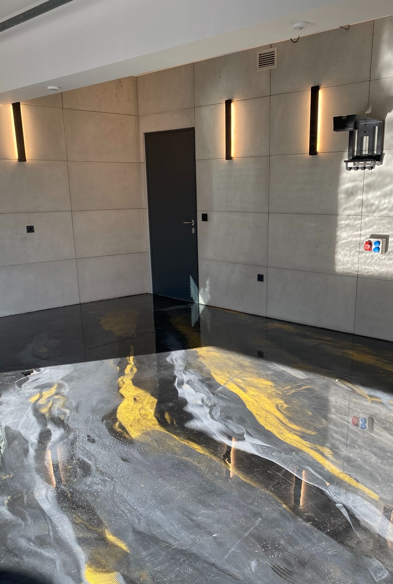 Błyszcząca, czarno-szaro-złota posadzka z żywicy w nowoczesnym wnętrzu z betonowymi płytami ściennymi i minimalistycznym oświetleniem. Widoczne odbicia światła.