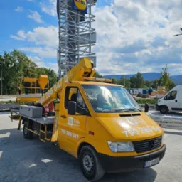 Żółty podnośnik koszowy z wysięgnikiem na tle konstrukcji reklamowej Redkom i Lidl, błękitne niebo z chmurami w tle, widok z dołu.