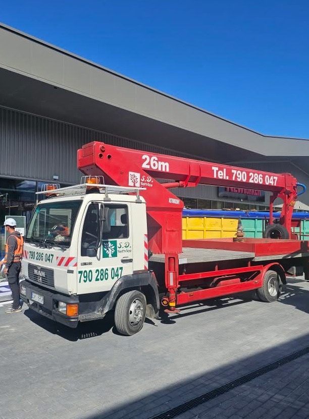 Czerwono-biały podnośnik koszowy MAN z wysięgnikiem 26m, logo S.D. Service i numerem telefonu, zaparkowany na placu z kostki, pracownik w odblaskowej kamizelce obok.