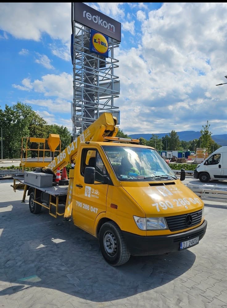 Żółty podnośnik koszowy z wysięgnikiem na tle konstrukcji reklamowej Redkom i Lidl, błękitne niebo z chmurami w tle, widok z dołu.