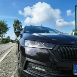 Ciemna Skoda Octavia, widok z przodu na tle błękitnego nieba z chmurami, zaparkowana na żwirowym poboczu drogi.