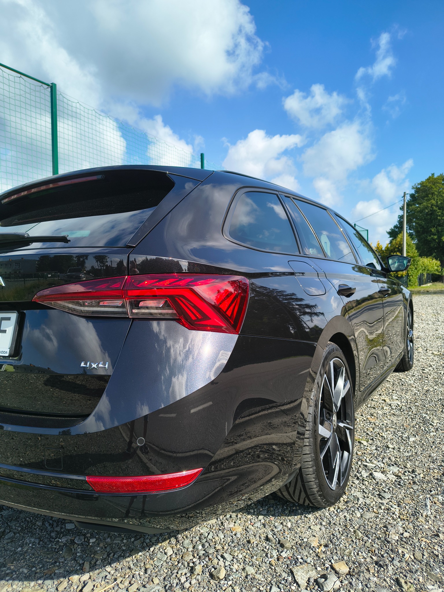 Błyszczący, czarny samochód typu kombi, Skoda Octavia, zaparkowany na żwirowym podłożu, odbijający błękitne niebo z chmurami. Widoczny fragment tylnej lampy i opony.