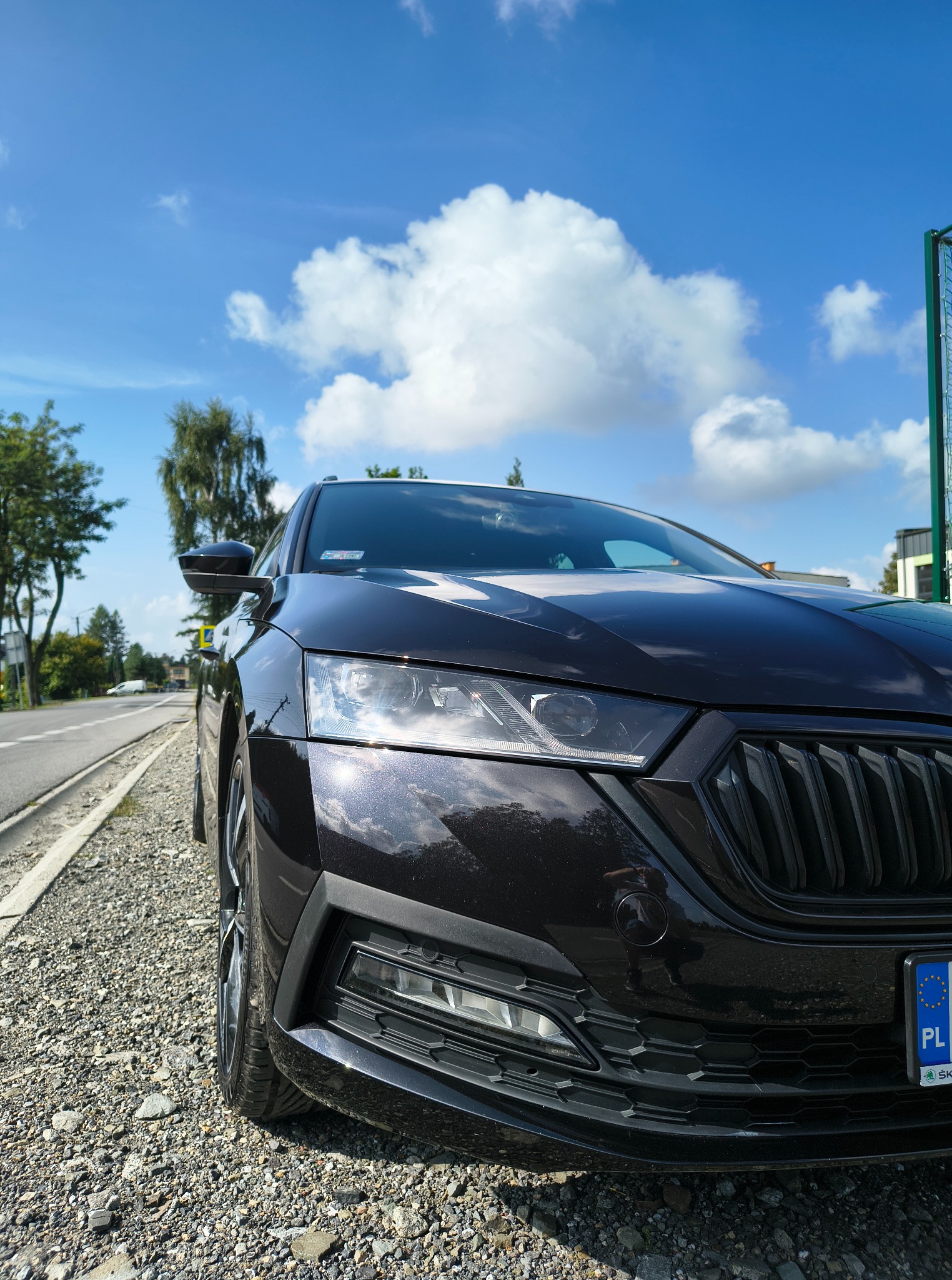 Ciemna Skoda Octavia, widok z przodu na tle błękitnego nieba z chmurami, zaparkowana na żwirowym poboczu drogi.