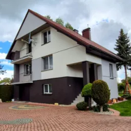 Dwukondygnacyjny dom z balkonami, pomalowany w odcieniach szarości i brązu, z widoczną anteną satelitarną na elewacji i ozdobnym brukiem na podjeździe.