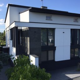 Tynqme Sp. z o.o. - Nowoczesny dom z białą elewacją i czarnymi akcentami, widok z zewnątrz w słoneczny dzień. Minimalistyczny design, duże okna, zadbany ogród.